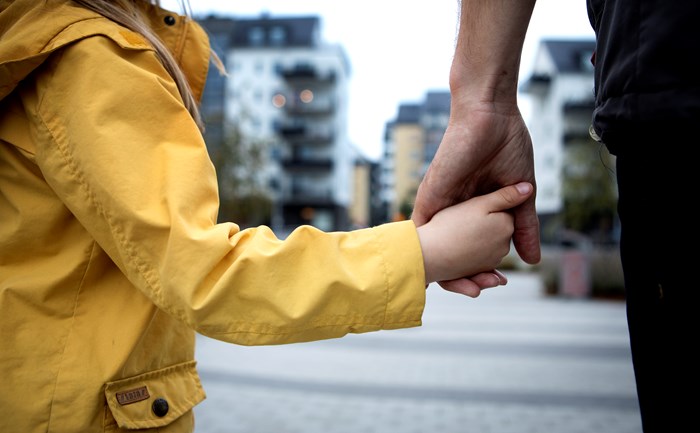 Närbild på en vuxen och ett barn som håller varandra i handen, fokus på händerna och ett bostadsområde med lägenheter i bakgrunden.