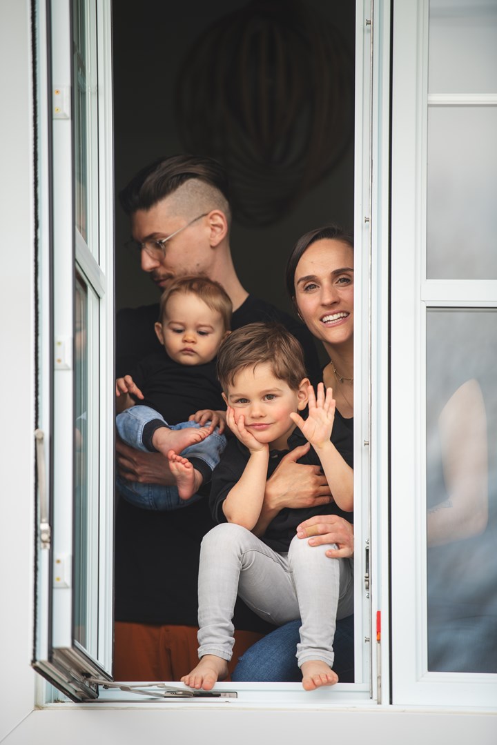 Familj med två vuxna och två barn kikar ut genom fönstret i sin lägenhet och vinkar.